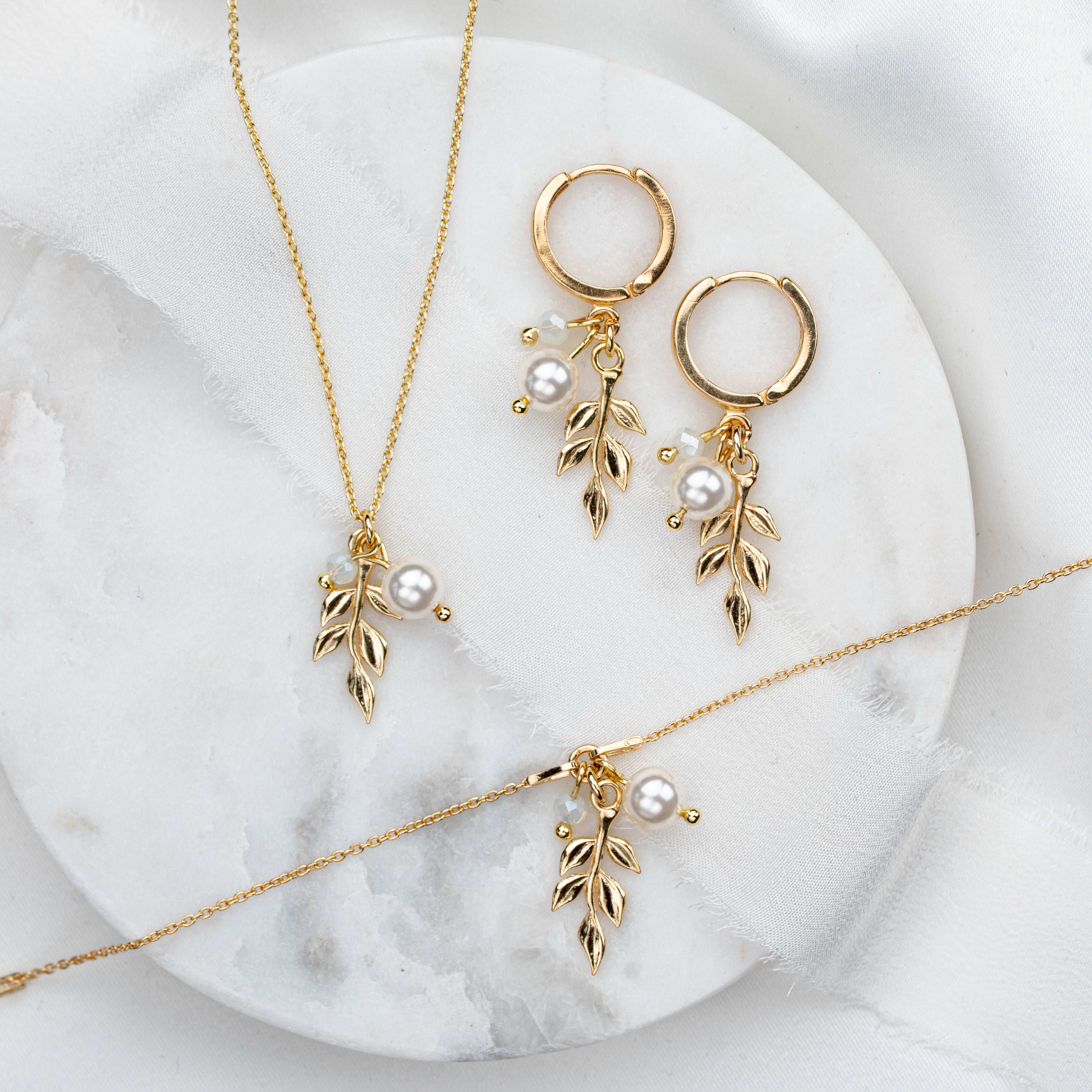 3-teiliges Schmuckset in 925er Silber 18K vergoldet bestehend aus Halskette, Creolen und Armband mit filigranen Blattanhängern, kleinen weißen Perlen und white opal Kristallperlen arrangiert auf hellem Marmorhintergrund