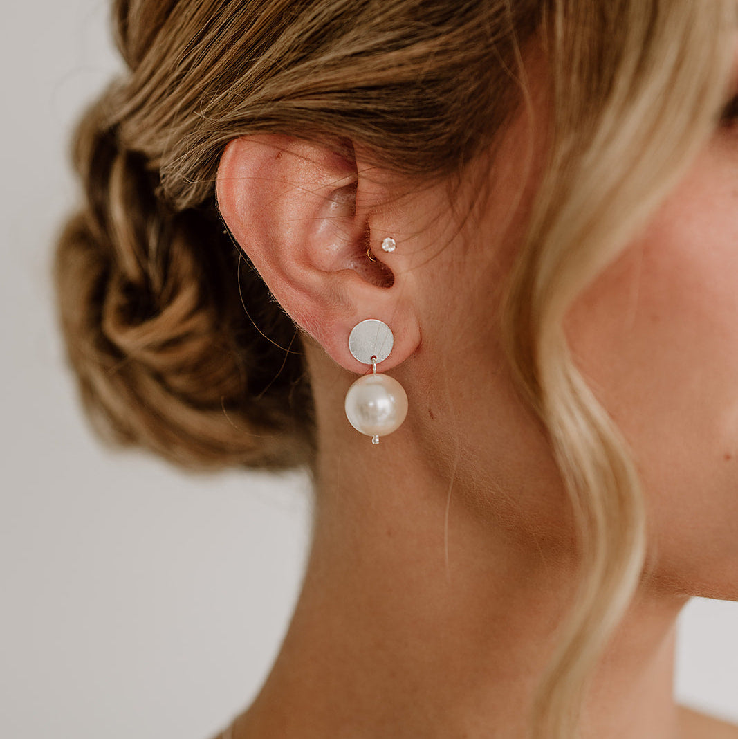 Zarte Brautohrringe aus 925er Sterlingsilber mit runder veganer Perle unter einem schlichten, runden Plättchen Ohrstecker, elegant getragen von einem Model am Ohr und harmonisch kombiniert mit einer Brautfrisur