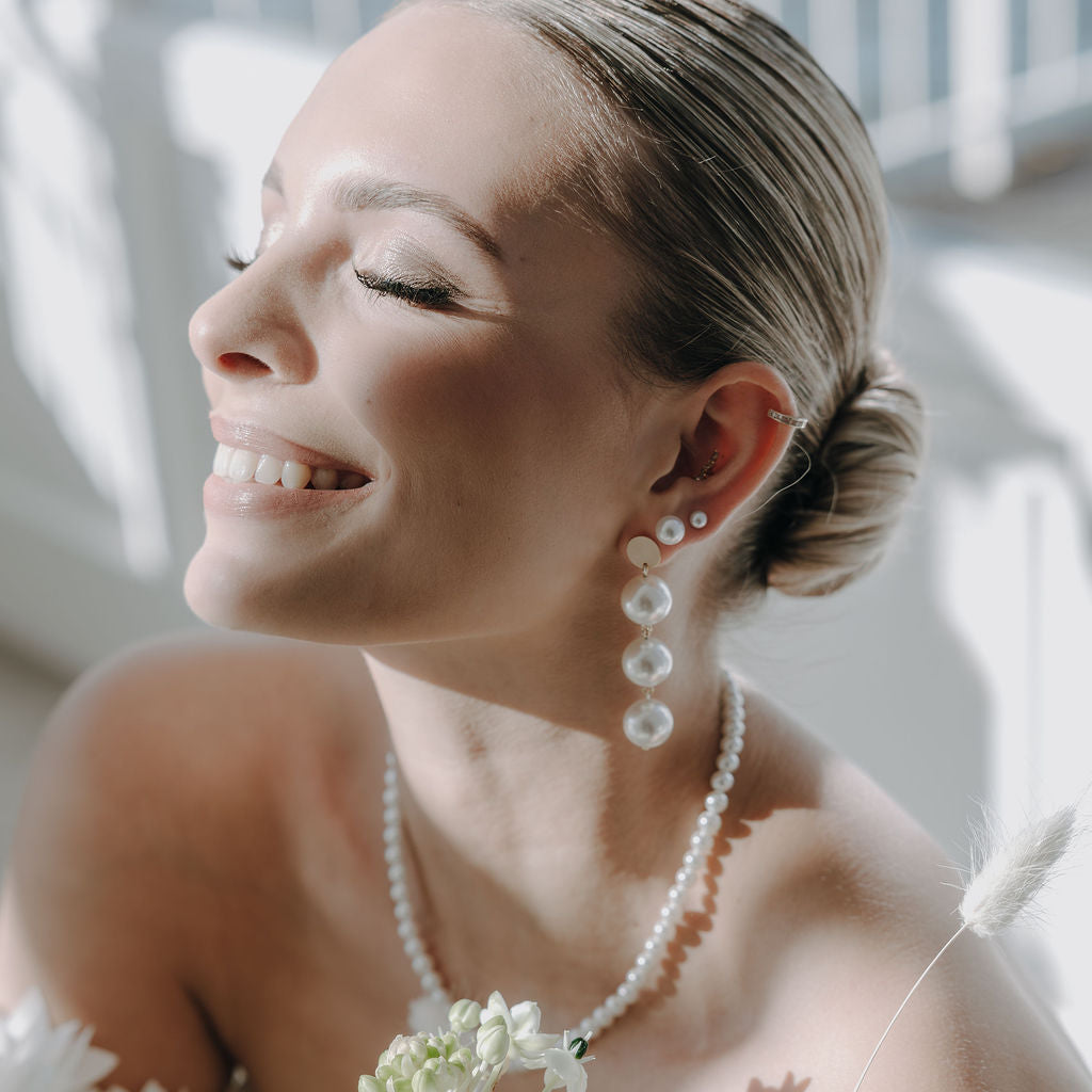 Elegante Brautohrringe aus 925er Sterlingsilber 18K vergoldet mit großen runden veganen Perlen untereinander hängend an einem schlichten, runden Plättchen Ohrstecker, stilvoll getragen von einem Model am Ohr und harmonisch kombiniert mit einer klassischen Perlenkette und einer eleganten Brautfrisur.