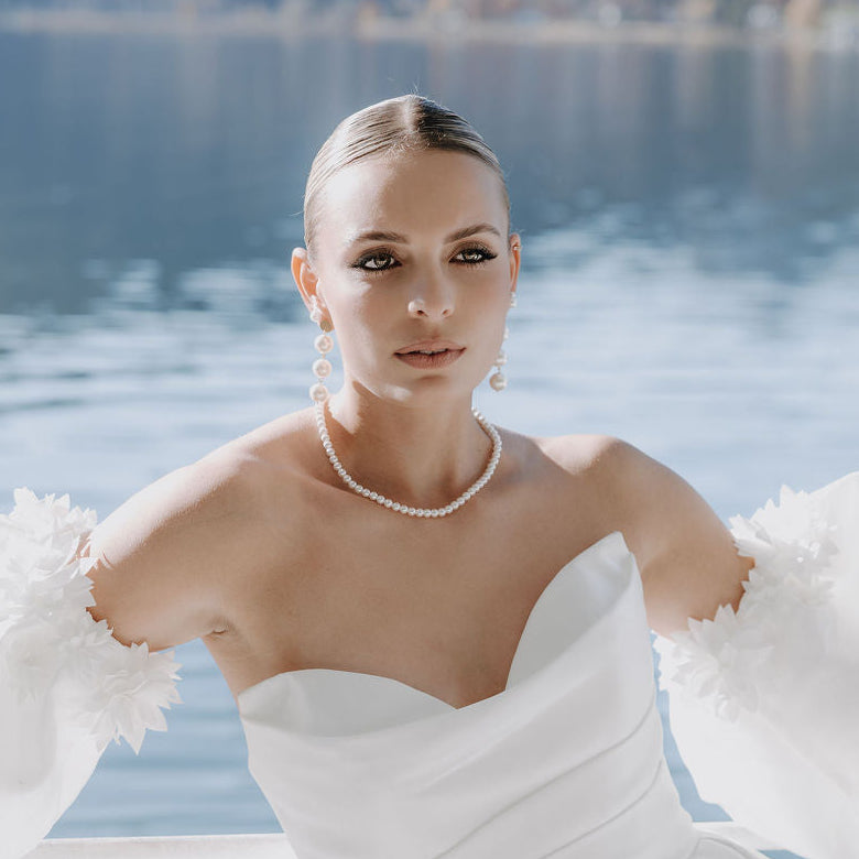 Elegante Brautohrringe aus 925er Sterlingsilber 18K vergoldet mit großen runden veganen Perlen untereinander hängend an einem schlichten, runden Plättchen Ohrstecker, stilvoll getragen von einem Model am Ohr und harmonisch kombiniert mit einer klassischen Perlenkette und einem schulterfreien Brautkleid.