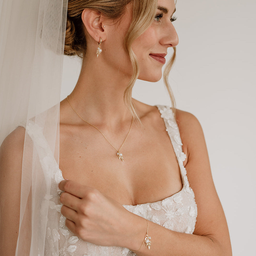 Model im weißen Brautkleid mit Schleier und elegantem Hochsteckfrisur-Look, trägt 3-teiliges Brautschmuckset in 925er Silber 18K vergoldet bestehend aus Halskette, Creolen und Armband mit filigranen Blattanhängern, kleinen weißen Perlen und white opal Kristallperlen.