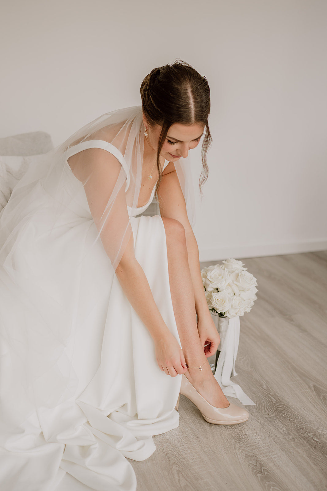 Braut bindet sich ein zartes Fußkettchen mit blauem Kristall an den Knöchel – detailreiche Getting-Ready-Szene mit Brautkleid und weißen Blumen.