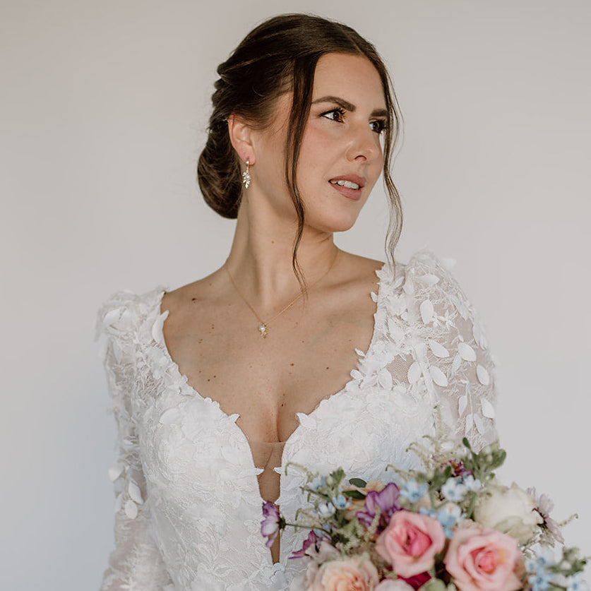 Model im weißen Brautkleid mit elegantem Hochsteckfrisur-Look, trägt ein Schmuckset in 925er Silber 18K vergoldet bestehend aus Halskette und Creolen mit filigranen Blattanhängern, kleinen weißen Perlen und white opal Kristallperlen.