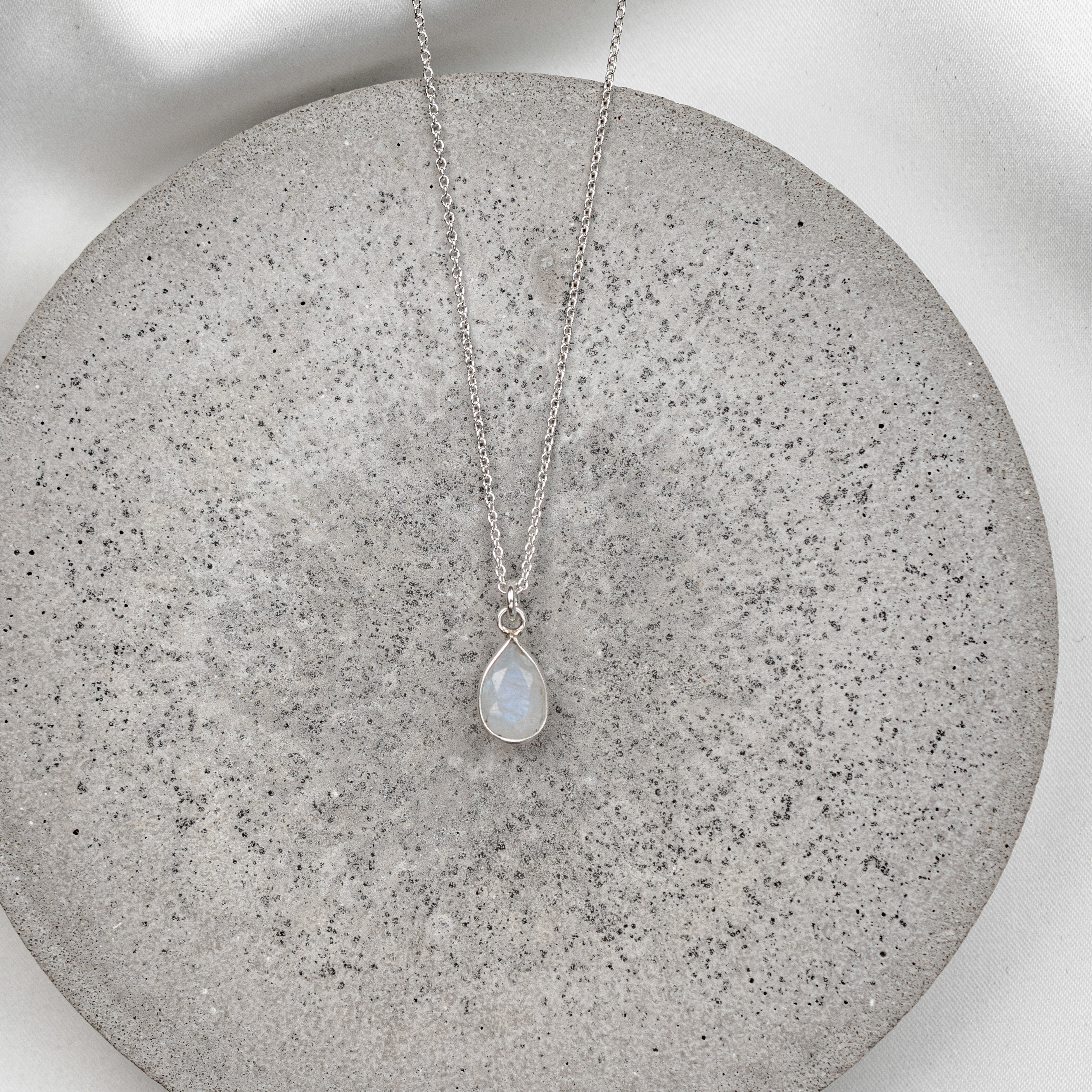 Braut Halskette aus 925er Sterlingsilber mit feiner Gliederkette und tropfenförmigem Mondstein Anhänger, präsentiert auf einer runden, hellgrauen Betonplatte