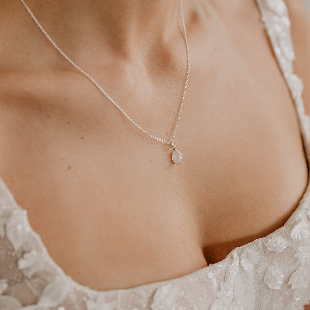 Zarte Brauthalskette aus 925er Sterlingsilber mit kleinem tropfenförmigem Mondstein Anhänger, getragen von einem Model auf dem Dekolleté, kombiniert mit einem weißen, spitzenbesetzten Brautkleid.