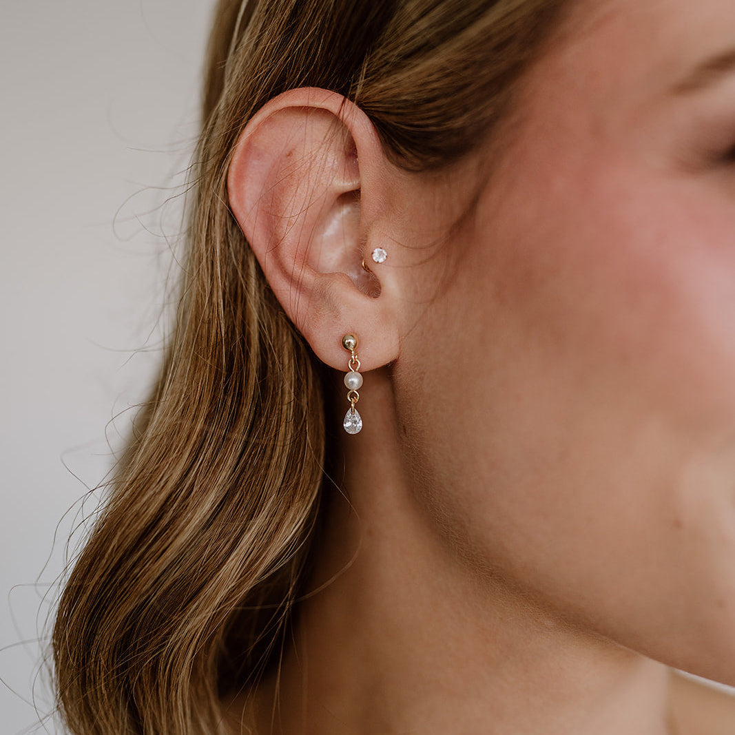 Zarte Brautohrringe aus 925er Sterlingsilber 18K vergoldet mit hängendem tropfenförmigem Zirkonia Kristall und kleiner runder Perle, elegant getragen von einem Model am Ohr und harmonisch kombiniert mit einer offenen Brautfrisur.
