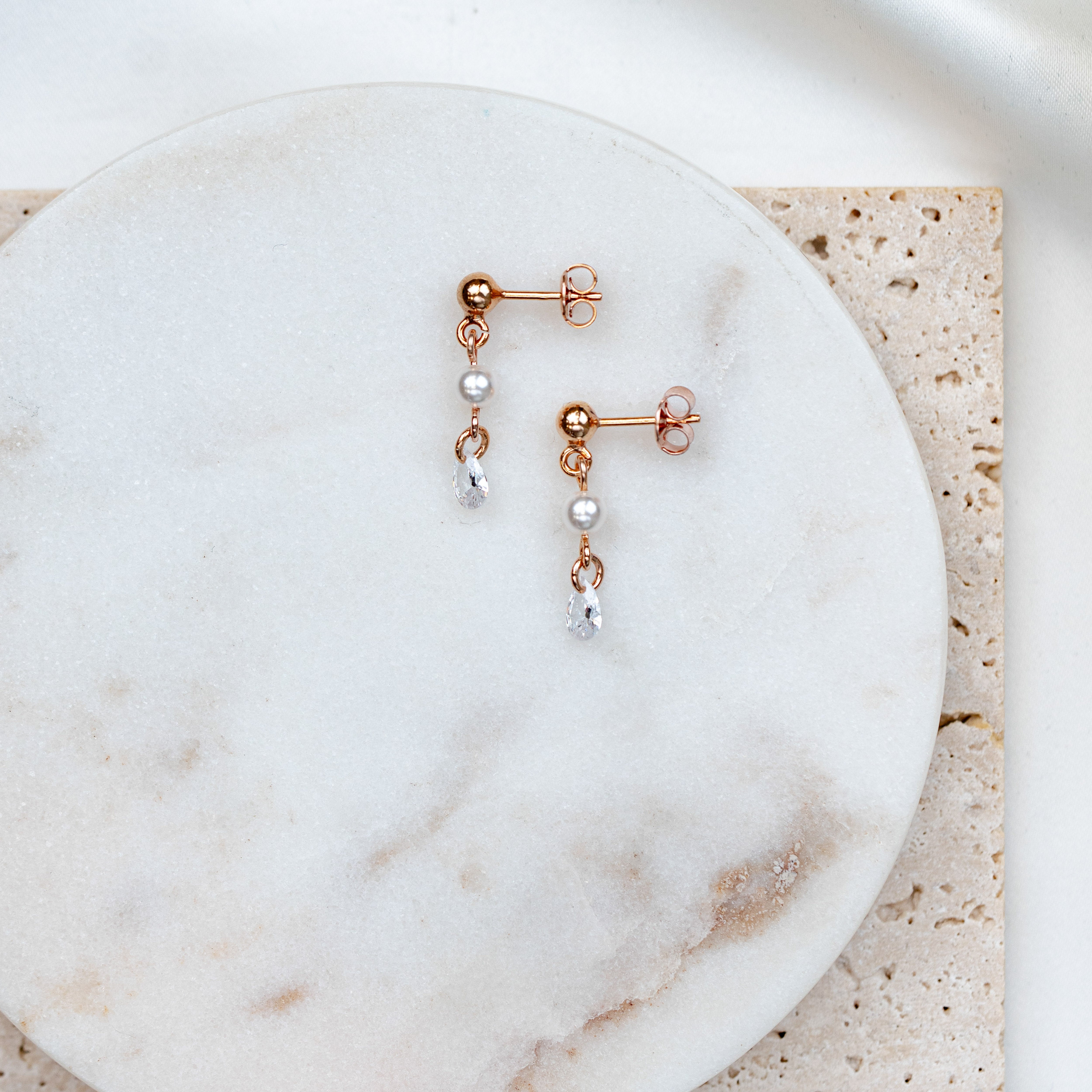 Zarte Brautohrringe aus 925er Sterlingsilber 18K rosévergoldet mit hängendem tropfenförmigem Zirkonia Kristall und kleiner runder Perler, stilvoll arrangiert auf einer hellen Marmorplatte.