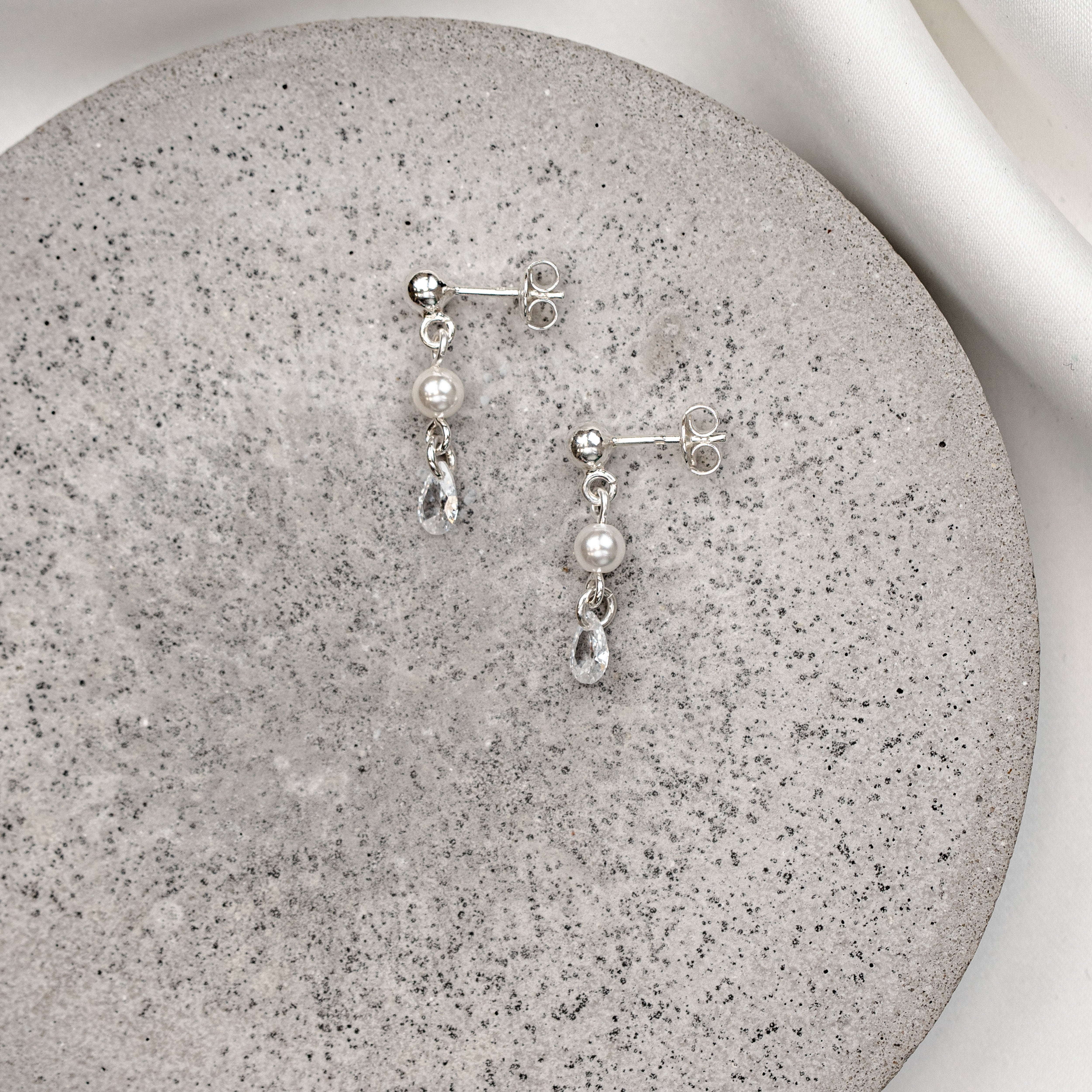 Zarte Brautohrringe aus 925er Sterlingsilber mit hängendem tropfenförmigem Zirkonia Kristall und kleiner runder Perle, stilvoll arrangiert auf einer hellgrauen, runden Betonplatte.