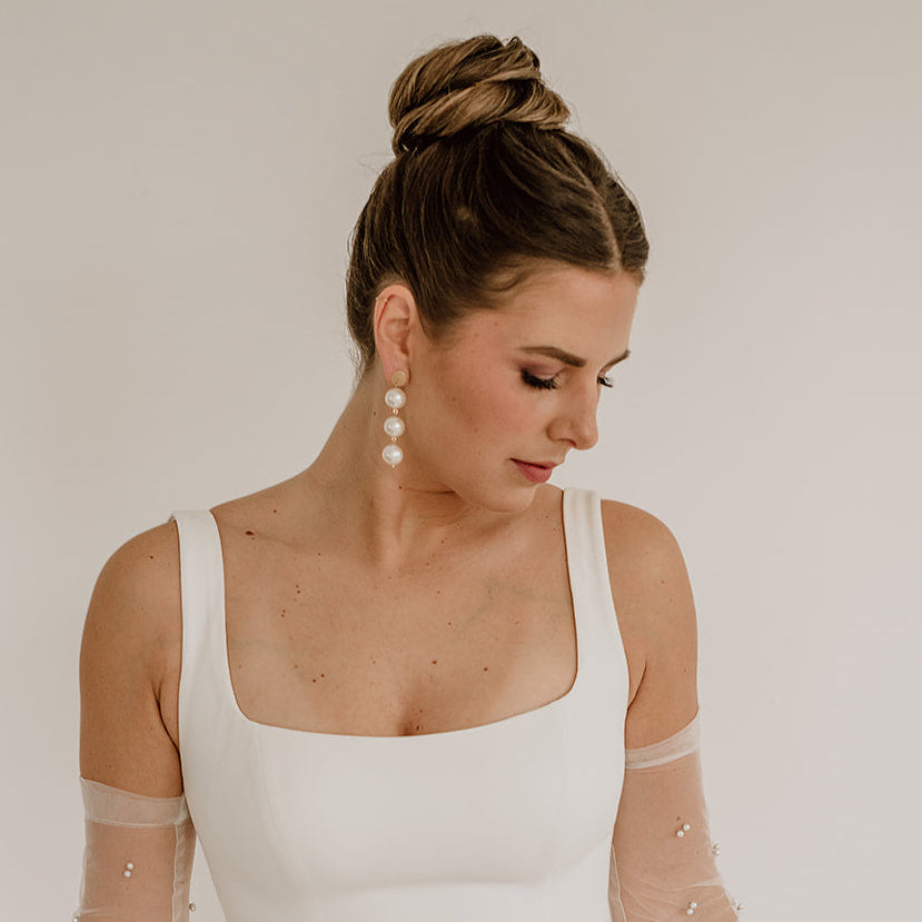 Elegante Brautohrringe aus 925er Sterlingsilber 18K vergoldet mit runden veganen Perlen untereinander hängend an einem schlichten, runden Plättchen Ohrstecker, stilvoll getragen von einem Model am Ohr und harmonisch kombiniert mit einem minimalistischen Brautkleid und einer eleganten Hochsteckfrisur.