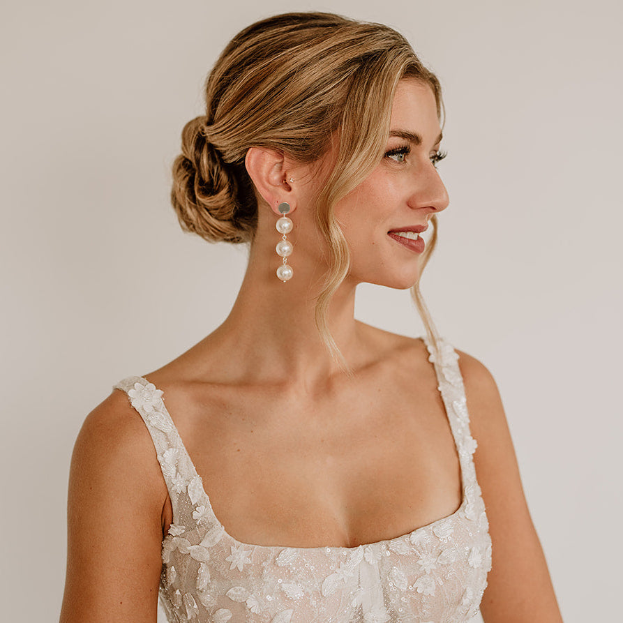 Elegante Brautohrringe aus 925er Sterlingsilber 18K vergoldet mit runden veganen Perlen untereinander hängend an einem schlichten, runden Plättchen Ohrstecker, stilvoll getragen von einem Model am Ohr und harmonisch kombiniert mit einem weißen, floral verzierten Brautkleid und einer eleganten Hochsteckbrautfrisur.