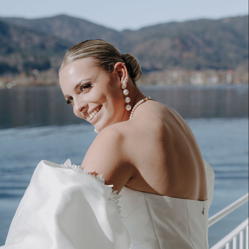 Elegante Brautohrringe aus 925er Sterlingsilber 18K vergoldet mit runden veganen Perlen untereinander hängend an einem schlichten, runden Plättchen Ohrstecker, stilvoll getragen von einem Model am Ohr und harmonisch kombiniert mit einer klassischen Perlenkette und einem schulterfreien Brautkleid.